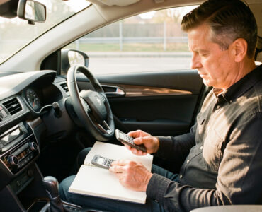 Un homme d’âge mûr calcule son budget automobile dans une voiture moderne, avec un carnet aux pages vierges et une calculatrice, éclairé par la lumière naturelle.